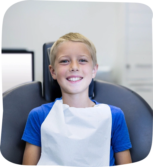 Young boy smiling in dental chair wearing blue and white shirt at pediatric dental office.